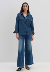 Camicia in denim con due tasche sul petto, maniche lunghe e chiusura con bottoni, abbinata a jeans azzurri a gamba larga e sandali neri.