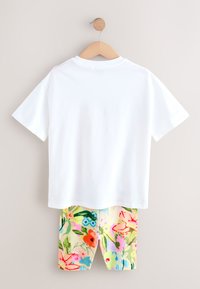 Wit katoenen T-shirt met korte mouwen hangt boven een korte broek met bloemenprint in levendige kleuren zoals roze, blauw en groen op een lichte achtergrond.