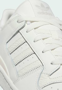 Close-up de um sneaker Adidas branco mostrando as riscas em pele texturizada, os atacadores e o logo da Adidas na lingueta.