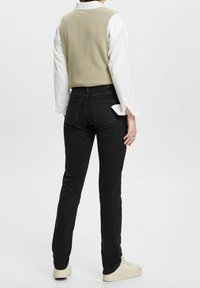 Vaqueros negros ajustados con cintura alta y bolsillos traseros, combinados con un chaleco de punto verde claro sobre una camisa blanca.