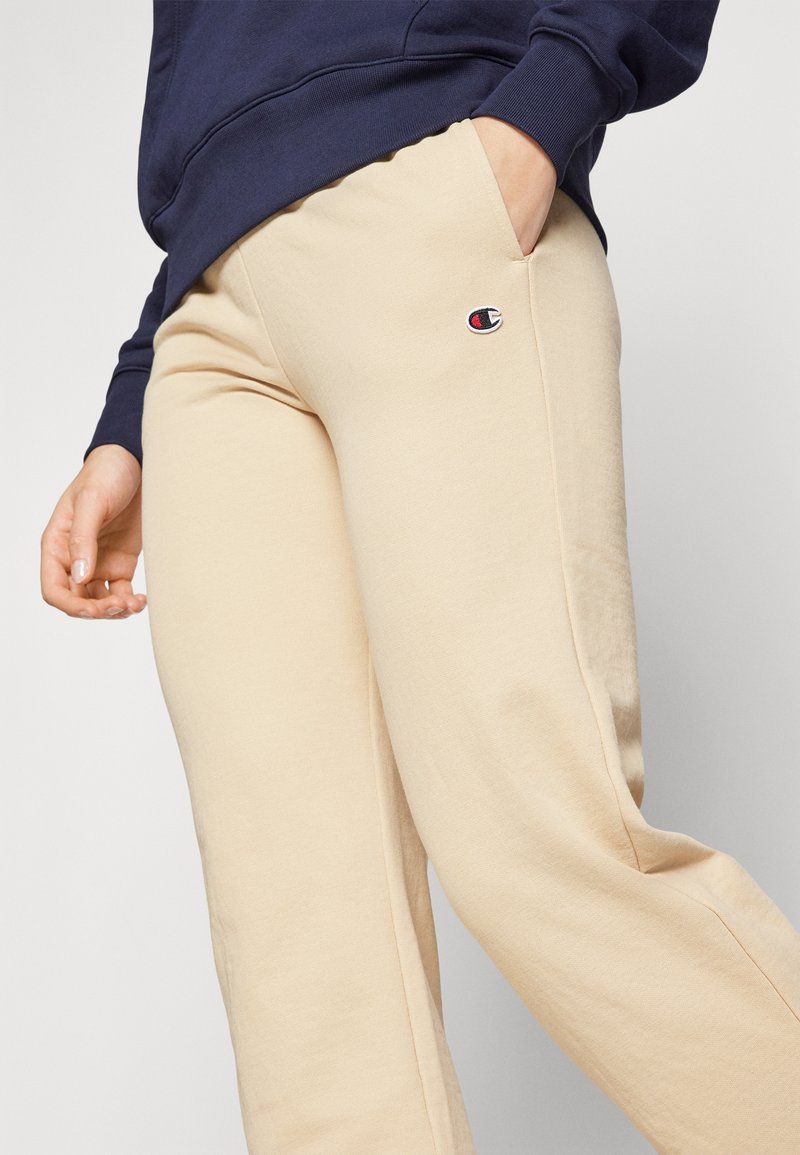 Pantalons de survêtement en coton beige avec une coupe décontractée, dotés de poches latérales et d'un petit logo brodé sur la cuisse. Texture douce et finition lisse.