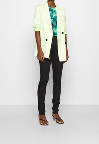 Blazer oversized verde chiaro con maniche arrotolate, abbinato a una camicia con motivo palme, pantaloni skinny neri e sandali con tacco neri.