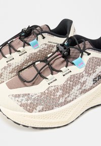 Paire de baskets de sport beiges et marron à motifs, lacets noirs à serrage rapide, œillets bleus et roses et semelle crème texturée.