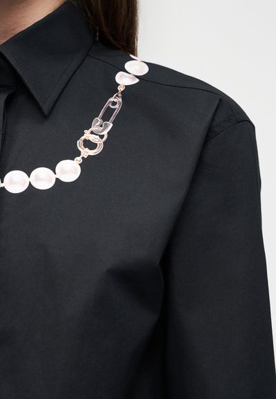 Μαύρο πουκάμισο με γιακά και κολιέ από πέρλες με κούμπωμα χρυσή παραμάνα, το οποίο ακουμπά στην περιοχή του ώμου.