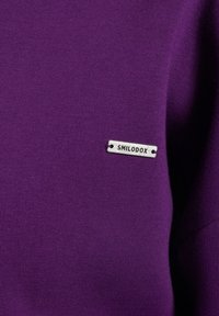 Suéter de algodón morado con una pequeña etiqueta plateada que dice "SMILODOX", situada en el lado izquierdo cerca del cuello.