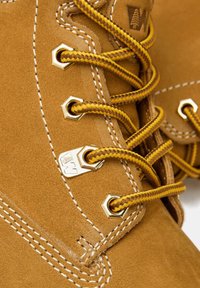 Stivale in suede marrone con occhielli dorati, lacci intrecciati gialli e cuciture bianche. Presenta una superficie testurizzata e un piccolo dettaglio del logo.