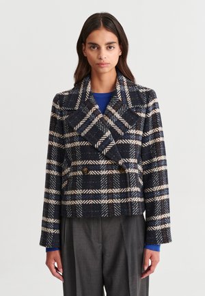 Femme portant une veste croisée à carreaux bleu marine, blanc et beige avec de larges revers, sur un haut bleu et un pantalon gris.