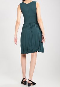 Robe sans manches teal avec un corsage ajusté et une taille froncée. Tissu lisse avec une fermeture éclair discrète au dos. Portée avec des sandales à talons noires.