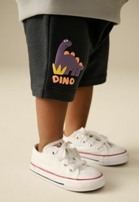Shorts noirs avec un graphique de dinosaure violet et le texte "DINO" en orange, associés à des baskets en toile blanches avec des accents rouges et bleus.