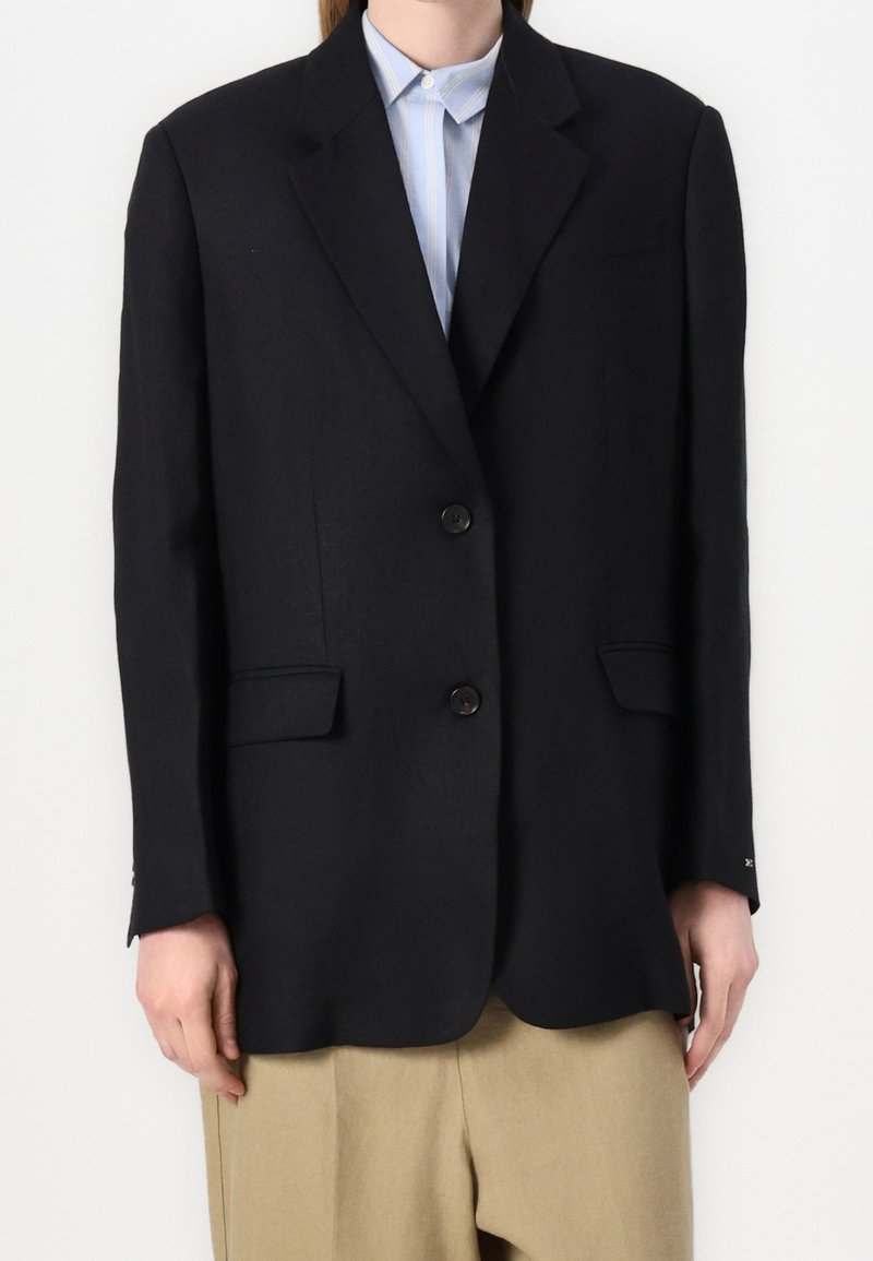 Personne portant un blazer noir à deux boutons sur une chemise bleu clair à col et un pantalon beige sur un fond blanc uni.