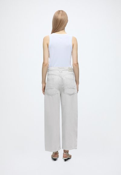 Femme aux cheveux blonds lisses porte un top blanc sans manches, un jean gris clair à jambes larges et des sandales, debout de dos à la caméra.