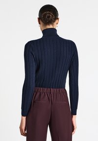 Maglione blu navy a collo alto con design a treccia a coste, abbinato a pantaloni bordeaux con vita elastica. La vista posteriore mette in risalto la texture e la vestibilità.