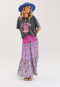 Šedé grafické tričko s designem růžové boty, spárované s maxi sukní s volány v růžových a modrých paisley vzorech. Světle hnělé obuv.