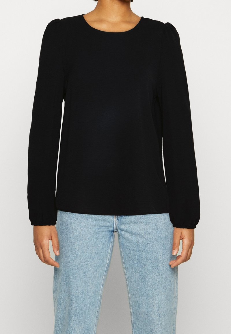 Personne portant un chemisier noir à manches longues avec des épaules bouffantes et un jean en denim bleu clair, debout, bras détendus le long du corps.