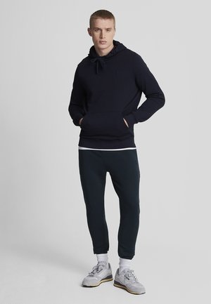 Marineblauwe hoodie met trekkoordcapuchon, buidelzak en logo; gecombineerd met donkere sweatpants en lichtgekleurde sportschoenen.
