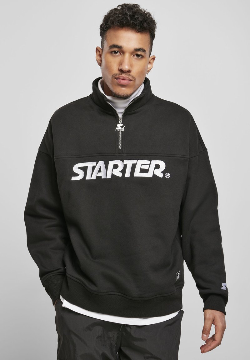 Svart hettegenser laget av teksturert stoff, med en halv glidelås-kollaro og stor hvit "STARTER" logo på brystet. Ermer og kant er ribbestrikket.