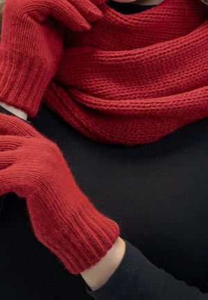 Personne portant des gants en tricot rouges et une écharpe assortie rouge par-dessus un haut noir, les bras croisés sur la poitrine.