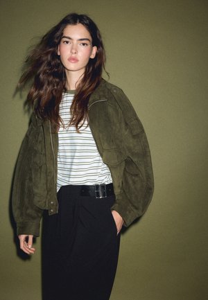 Jeune femme aux longs cheveux bruns portant une veste vert olive, une chemise blanche rayée et un pantalon noir sur fond vert.