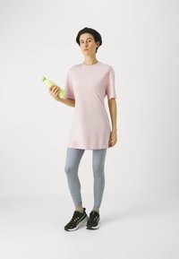 Svijetlo ružičasta oversized majica, sivi tajice, crne tenisice s zelenim akcentima, drži zelenu izoliranu bocu za vodu. Minimalistička pozadina.
