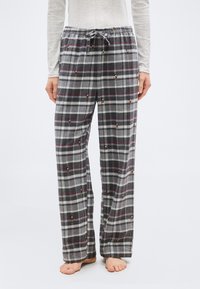 Personne portant un pantalon de pyjama gris à carreaux avec de petites imprimés Mickey Mouse et un haut à manches longues de couleur claire, debout pieds nus.