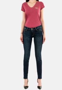 Camiseta rosa de cuello en V con mangas enrolladas, combinada con jeans ajustados azul oscuro que tienen detalles en dorado y costuras, llevada con tacones negros.