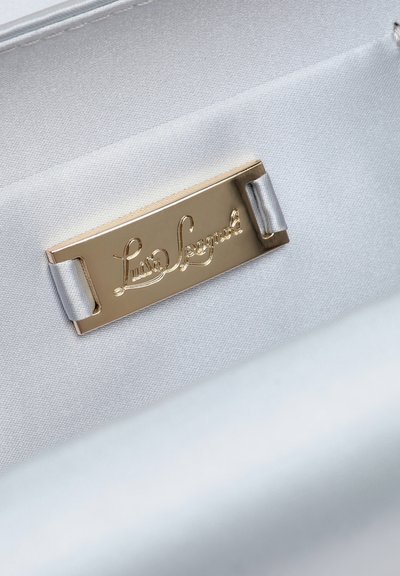 Metalkobberfarvet navneskilt med den graverede kursivtekst "Luisa Spagnoli" fastgjort til hvidt stof med sølvfarvede løkker.