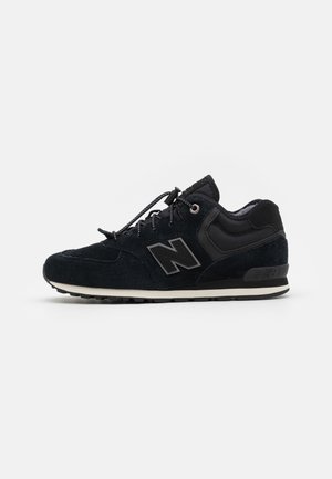 Chaussure de sport en suède noir et maille avec logo « N » réfléchissant, lacets élastiques à bouton de serrage et semelle rembourrée sur fond blanc.