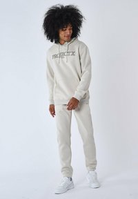 Sudadera beige con el texto "PROJECT X PARIS", bolsillo delantero y cordones blancos, combinada con joggers beige a juego y zapatillas blancas.