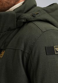 Green jacket featuring a textured fabric, zippered hood detail, and black hardware accents. Includes a brand patch on the shoulder.