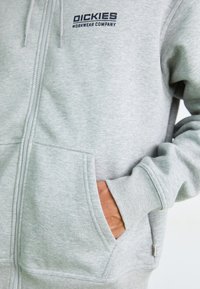 Hanorac gri cu fermoar, fabricat din material moale, având o pocket frontală, mansete ribbate și un logo negru "Dickies Workwear Company" pe pieptul stâng.