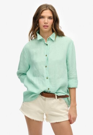 Chemise boutonnée vert clair avec une coupe décontractée, manches retroussées et boutons marron, associée à un short blanc déchiré et une ceinture marron.