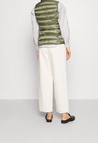 Oliwkowa kamizelka puchowa, widok od tyłu, z poziomym pikowaniem. W połączeniu z białą i szarą koszulą w paski oraz kremowymi spodniami o szerokich nogawkach.