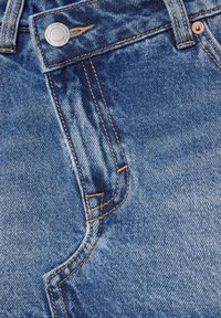 Gros plan sur un jean en denim bleu montrant la poche avant, la boucle de ceinture et un bouton métallique avec des détails de couture.