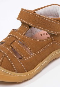 Ruskea nahkainen sandaali, jossa on ristikkäiset hihnat ja tarranauha. Vaalean värinen ommel ja kumipohja, jossa on karhea pinta.