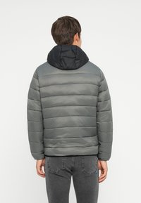 Grå pufferjakke med en sort hætte, der har vandrette quiltede paneler og en glat tekstur. Båret med grå jeans.