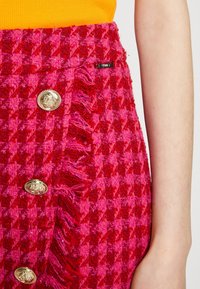 Jupe à carreaux rose et rouge en tissu texturé, avec des boutons dorés et des détails en franges le long de l'ourlet.