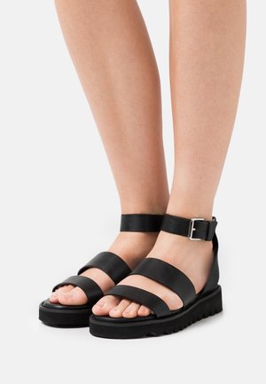 Pieds chaussés de sandales en cuir noir avec deux brides sur les orteils, une bride à la cheville avec une boucle argentée, et des semelles épaisses rainurées.