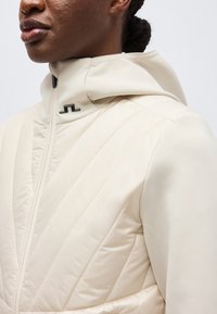 Közeli kép egy személyről, aki krémszínű, steppelt dzsekit visel, magas gallérral és fekete cipzárral, amely bemutatja a textúrát és egy kis fekete logót a galléron.