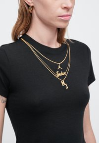 Złote naszyjniki warstwowe z zawieszkami z napisami "Jordan" i "MJ" ozdabiają czarną, dopasowaną koszulkę. Naszyjniki mają gładkie, błyszczące wykończenia.