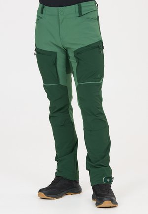 Mann trägt dunkel- und hellgrüne Outdoor-Hose mit Reißverschluss-Taschen am Oberschenkel und schwarze Wanderschuhe vor weißem Hintergrund.