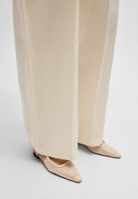 Płaskie buty w kształcie czubków w jasnobeżowej siateczce z metalicznymi akcentami. Noszone z luźnymi, kremowymi spodniami o szerokich nogawkach. Widać płaskie czarne podeszwy.