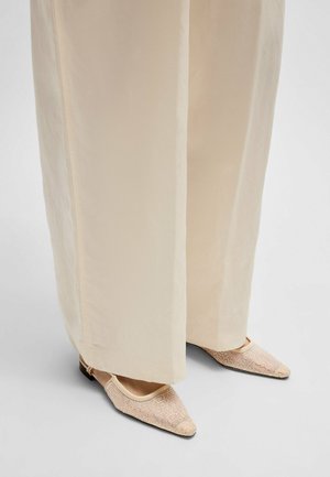 Lichte beige mesh ballerina's met metallic accentpatronen. Draagt met losse, crèmekleurige wijde broekspijpen. Platte zwarte zolen zichtbaar.