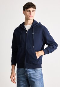 Hanorac cu fermoar de culoare bleumarin, fabricat dintr-un material moale, având o buzunar frontal, sfori pentru ajustare și broderie cu logo pe piept. Asortat cu blugi de culoare albastru deschis.