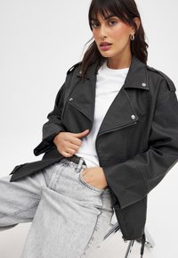 Schwarze Lederjacke mit Reverskragen, Reißverschlusstaschen und silbernem Zubehör, kombiniert mit einem weißen T-Shirt und hellgrauen, weit geschnittenen Jeans.