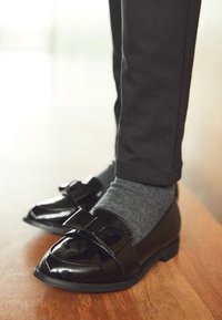 Sorte laklædersko med sløjfedetalje, flad sål og spids snude, kombineret med grå sokker og sorte, tætsiddende bukser.