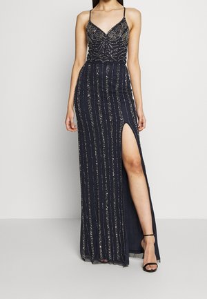 Frau trägt ein marineblaues Paillettenabendkleid mit dünnen Trägern und einem hohen seitlichen Schlitz, kombiniert mit schwarzen Knöchelriemen-Absätzen.