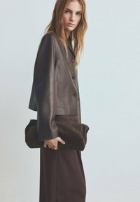 Brązowa krótka kurtka skórzana z kołnierzem i czterema guzikami, noszona z ciemnobrązową spódnicą, trzyma miękki brązowy zamszowy kopertówkę.