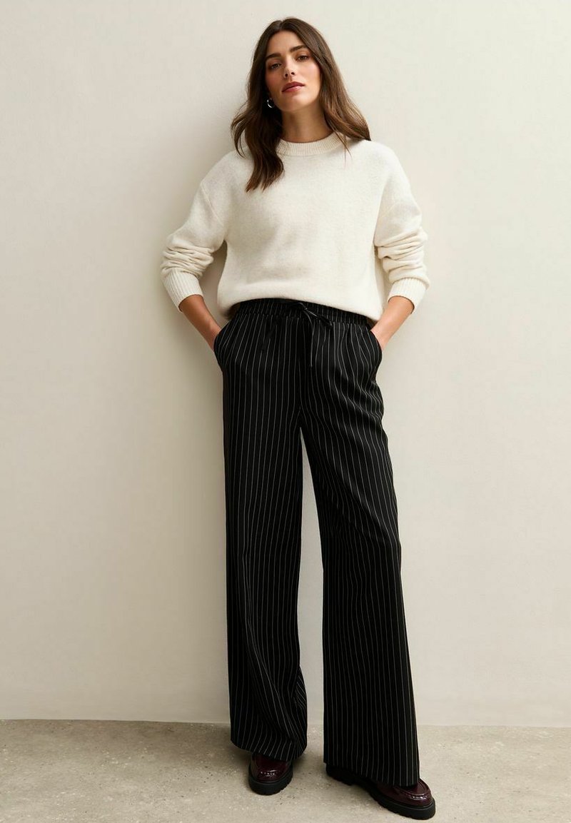 Suéter de gran tamaño en color crema con puños acanalados, combinado con pantalones de pierna ancha a rayas negras y zapatos de plataforma. Textura suave y corte relajado.