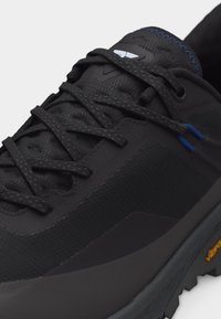 Κοντινή λήψη μαύρου πεζοπορικού παπουτσιού με υφασμάτινη υφή, μαύρα κορδόνια, λεπτομέρεια ραφής σε μπλε χρώμα και λογότυπο Vibram στη χοντρή σόλα.