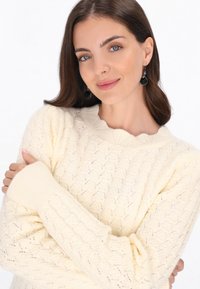 Krém színű, cable-knit pulóver fodros dekoltárral és hosszú bordás ujjakkal. A textúra gyémánt mintázatú. Fekete csepp fülbevalók láthatók.